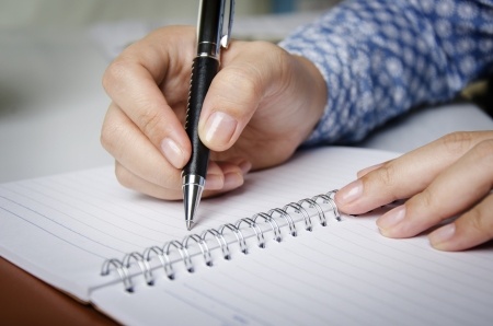 A hand holding a pen is shown writing in a notebook.