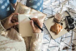 A woman sits near the camera, writing notes while enjoying breakfast food on the table.
