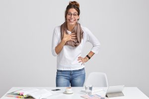 A smiling coach stands at her desk with one hand on her chest, exuding confidence and ease, embracing the idea of learning without fear of mistakes.
