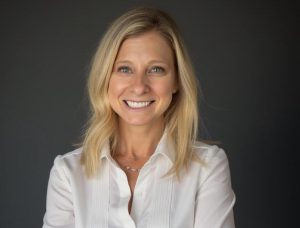 Ellen Rudofsky, PCC, CPCC smiling in a professional headshot, wearing a white button-up shirt against a dark gray background. She has shoulder-length blonde hair and a warm, approachable expression while facing the camera.