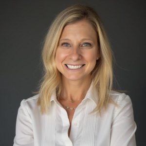 Ellen Rudofsky, PCC, CPCC smiling in a professional headshot, wearing a white button-up shirt against a dark gray background. She has shoulder-length blonde hair and a warm, approachable expression while facing the camera.